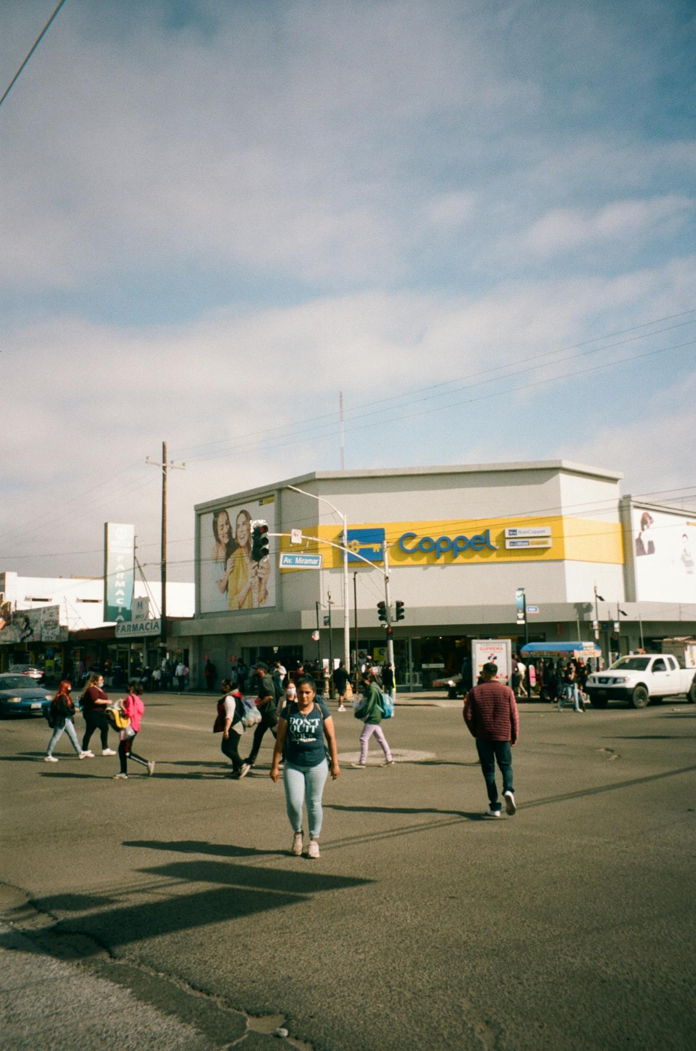 People crossing a vibrant street intersection in Ensenada, Mexico under a cloudy sky.